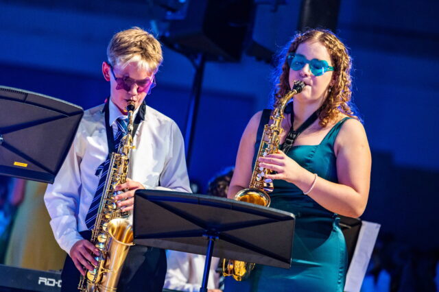 Two musicians wearing blue-tinted glasses play saxophones on stage, reading sheet music. The man wears a tie and shirt, the woman wears a teal dress. Blue lighting creates a vibrant atmosphere.