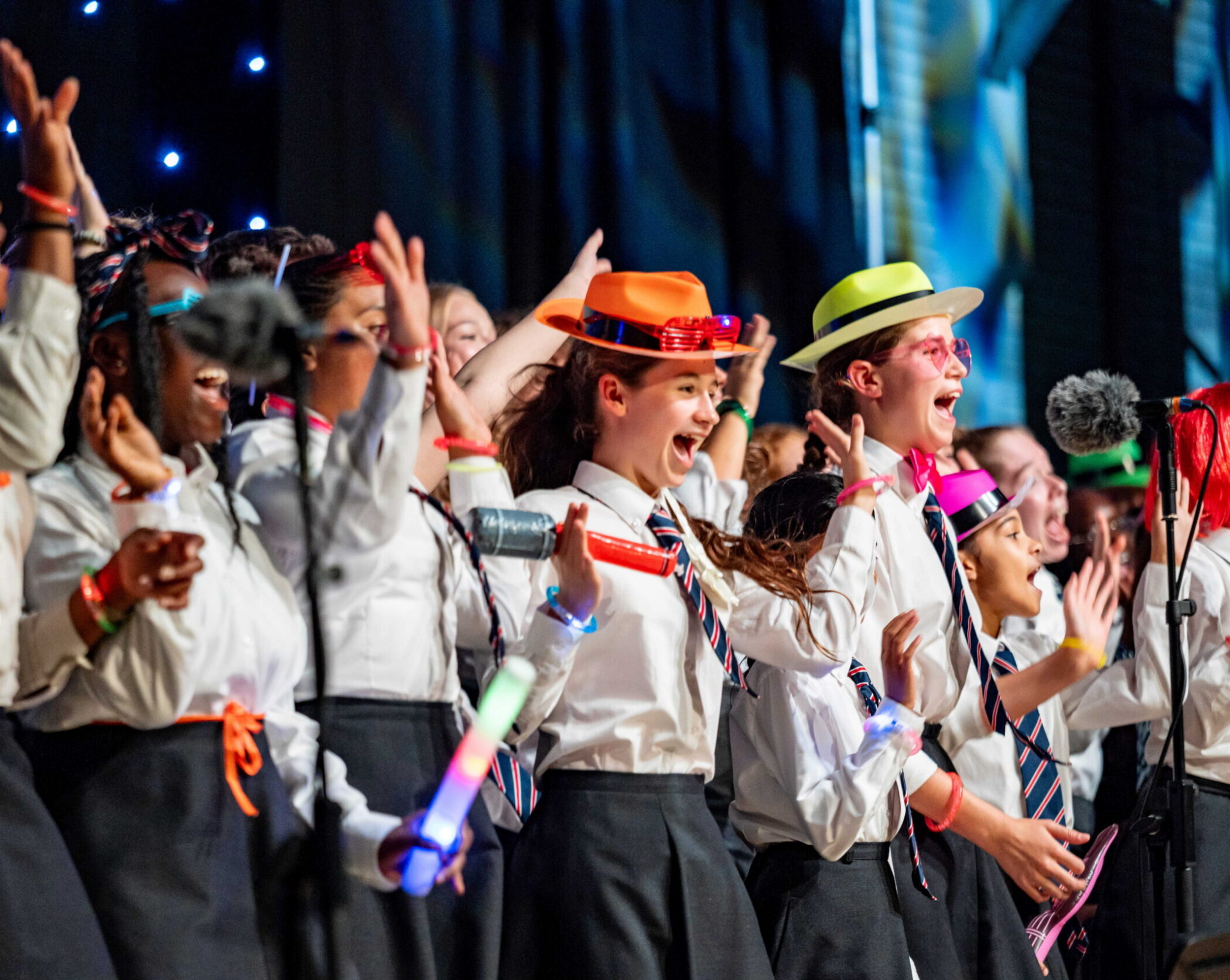A group of enthusiastic children in white shirts and ties sing on stage, some wearing colourful hats and holding glow sticks, with faces joyful and expressive under bright stage lights.