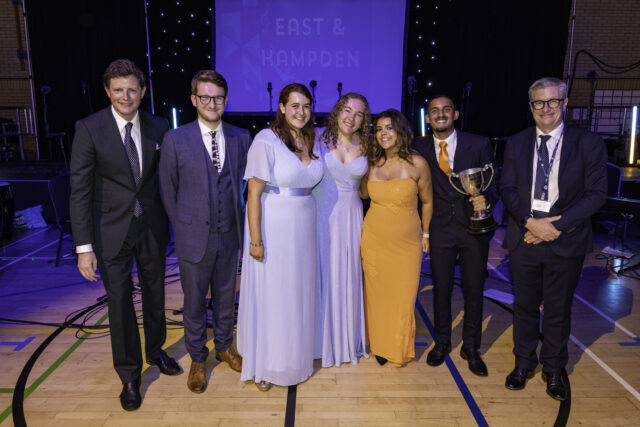 A group of seven people in formal attire stand smiling indoors on a sports hall floor. One person holds a trophy. Blue and orange dresses stand out. A stage with EAST & HAMPDEN signage is visible behind them.