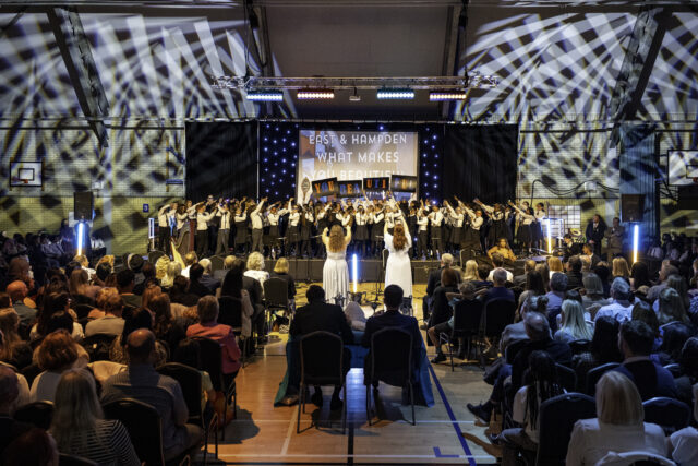 A choir of children sings on stage in front of a large audience in a gymnasium. Two adults in white dresses stand facing the group. Stage lights and a sign that reads, What makes you beautiful, are visible.