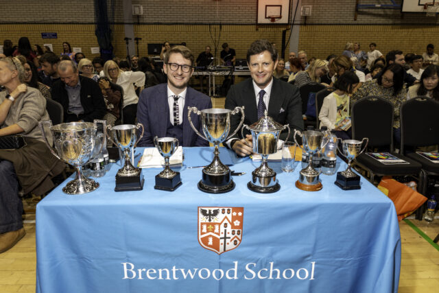 Two men in suits sit behind a table with trophies and a Brentwood School banner, facing an audience in a gymnasium. The audience appears engaged, with some people looking at the camera and others looking elsewhere.