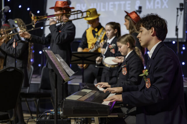 A group of students in uniforms perform on stage; one plays a keyboard while others play brass and string instruments, some wearing colourful hats. Blue stage lights twinkle in the background.