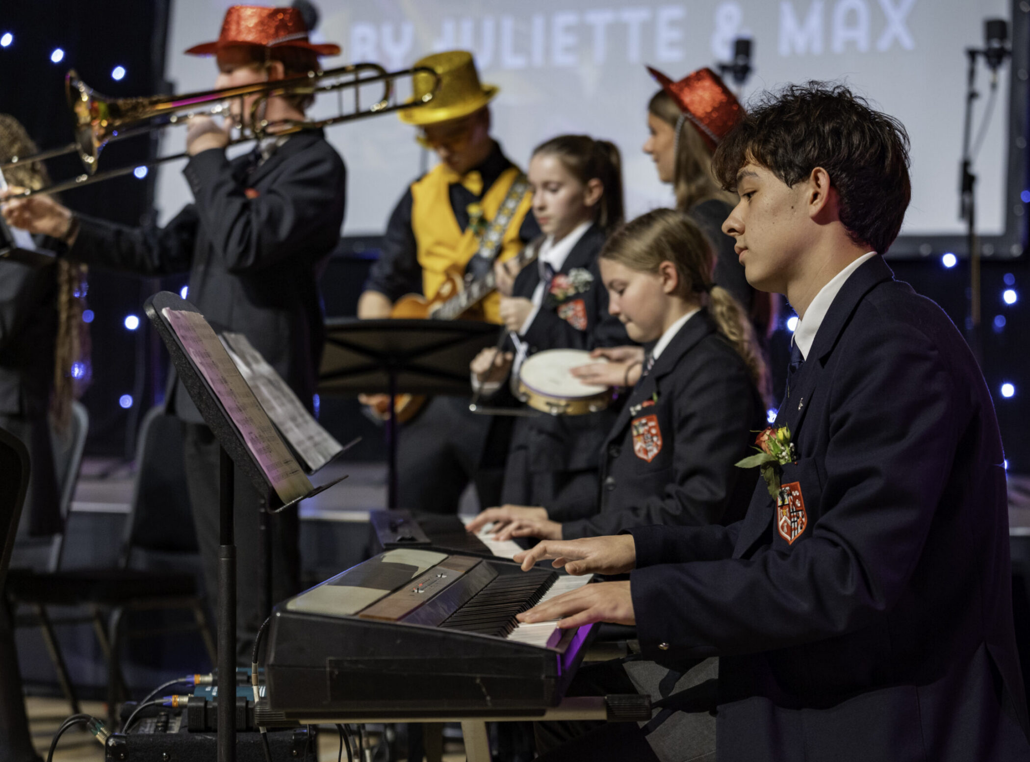 A group of students in uniforms perform on stage; one plays a keyboard while others play brass and string instruments, some wearing colourful hats. Blue stage lights twinkle in the background.