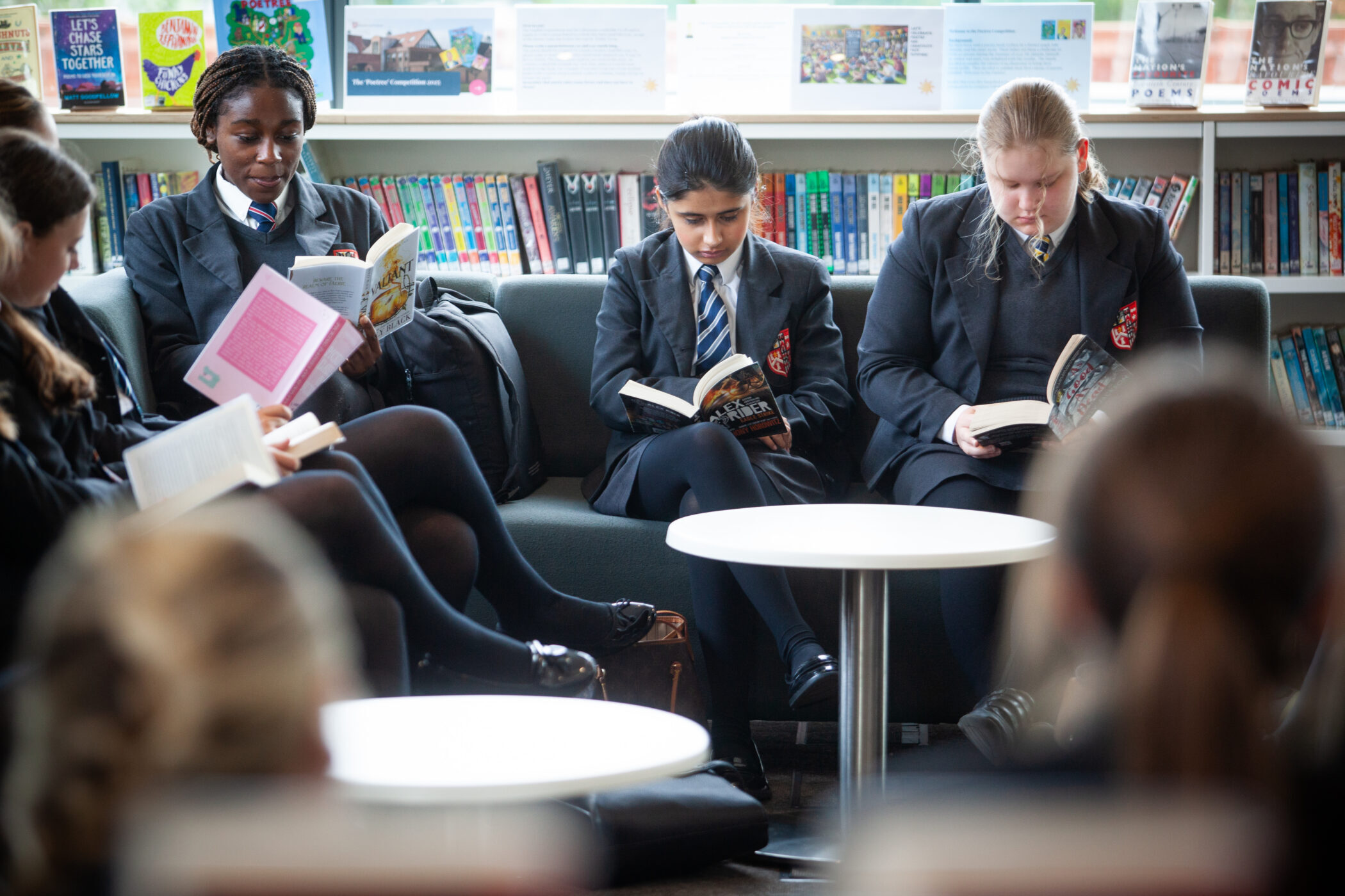 Three students in school uniforms sit on a sofa in a library, enjoying the excellent facilities as they read books. Shelves filled with books are visible in the background, and a white round table is in front of them.