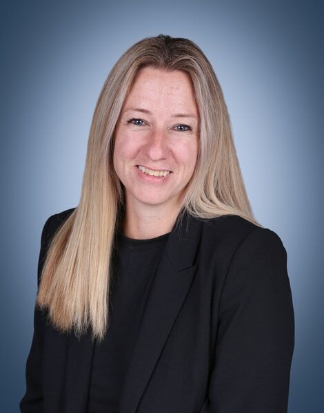 Smiling woman with long straight blonde hair, wearing a black blazer and black top, posed against a gradient blue background.