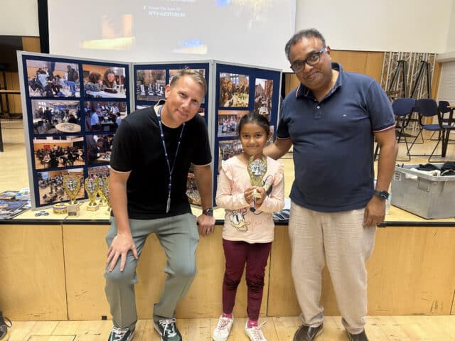 Three people stand indoors in front of a display board with photos. A young girl in the centre holds a trophy, smiling, while two adults stand beside her, also smiling. Trophies are visible on the table behind them.