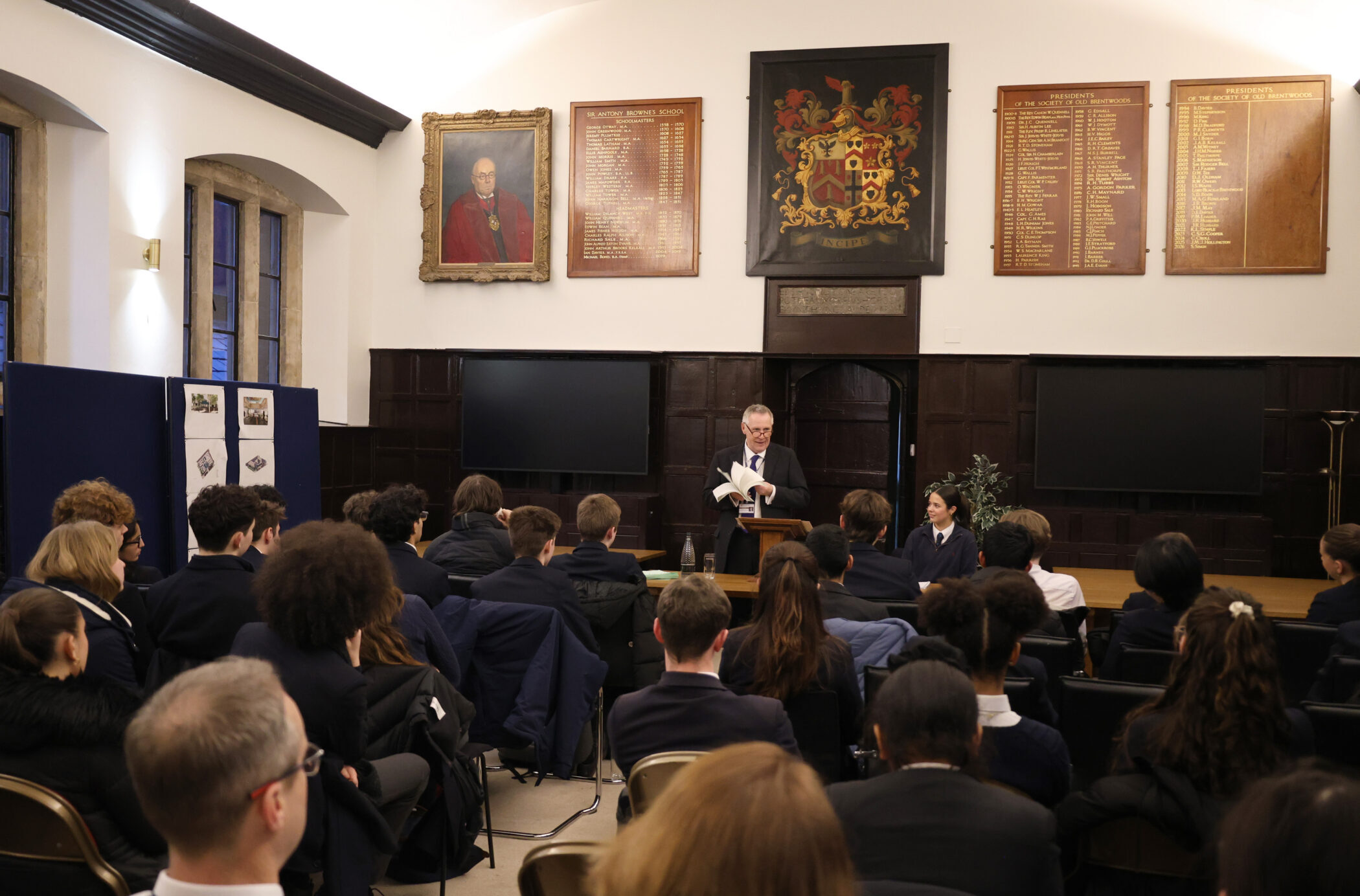 A man stands at a lectern reading to an audience seated in a formal room with wood panelling, portraits, and name plaques on the walls, one of the distinguished facilities designed for such gatherings. Most attendees are dressed in dark clothing.
