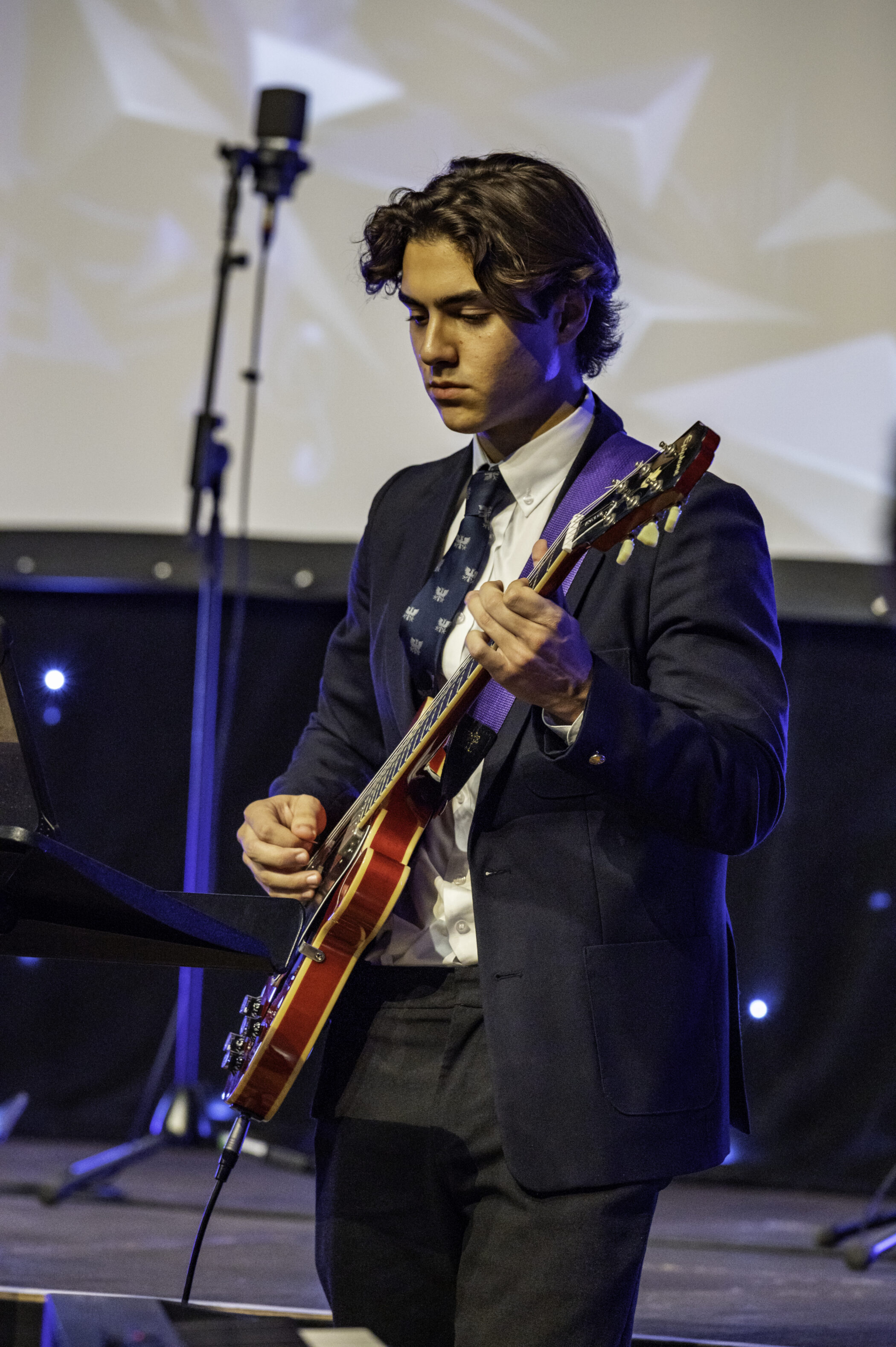 A young man in a suit, plays an electric guitar on stage, focusing intently. A microphone and stand are visible in the background, with blue lights and a blurred screen behind him.