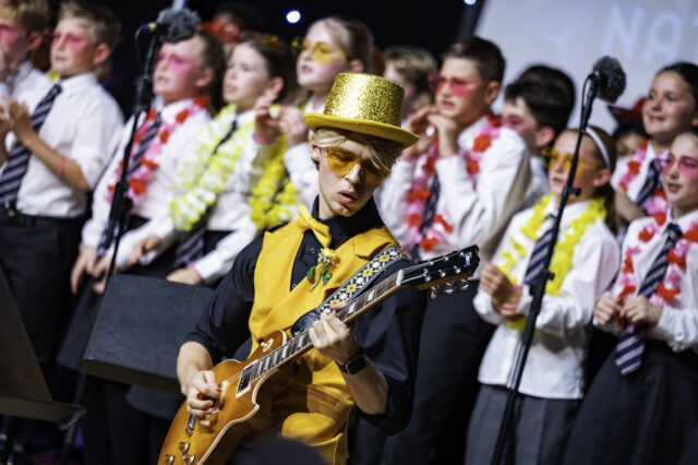 A person in a gold hat and waistcoat plays guitar on stage, while a group of children in white shirts, striped ties, and colourful leis sing and clap in the background.