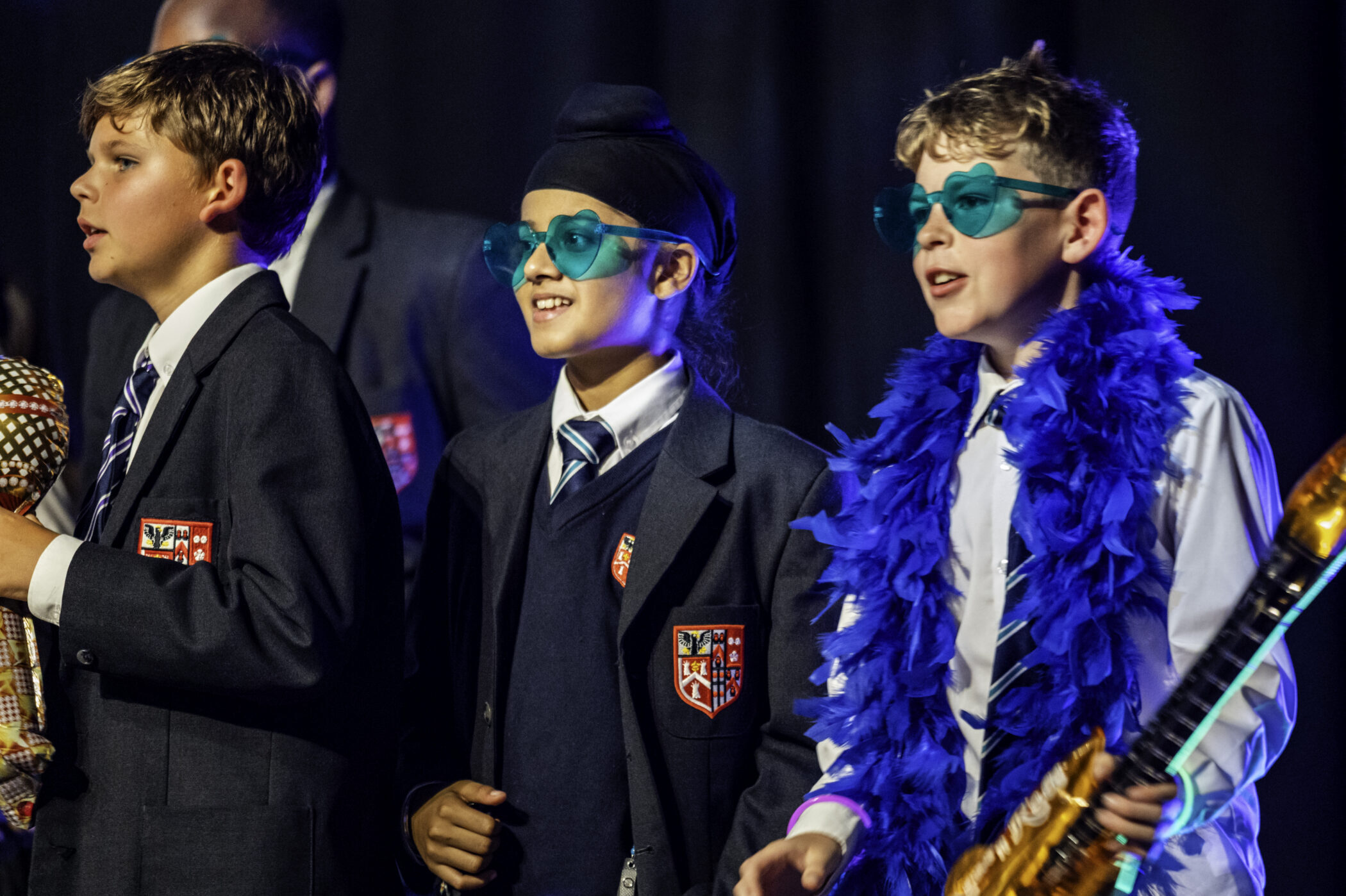 Three children in school uniforms, two wearing blue sunglasses and one with a blue feather boa, stand together smiling on a stage with a dark background.