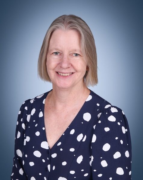 A woman with short, straight grey hair smiles at the camera. She is wearing a navy blue blouse with large white spots and is posed against a plain blue gradient background.