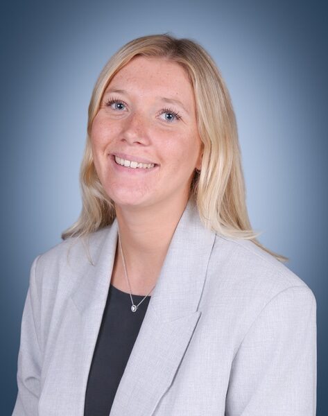 A woman with long blonde hair smiles, wearing a light grey blazer over a dark top, with a necklace. She is posed against a blue gradient background.