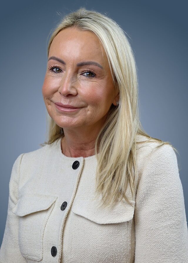 A woman with long blonde hair, wearing a cream-coloured textured jacket with black buttons, sits against a blue gradient background and smiles at the camera.