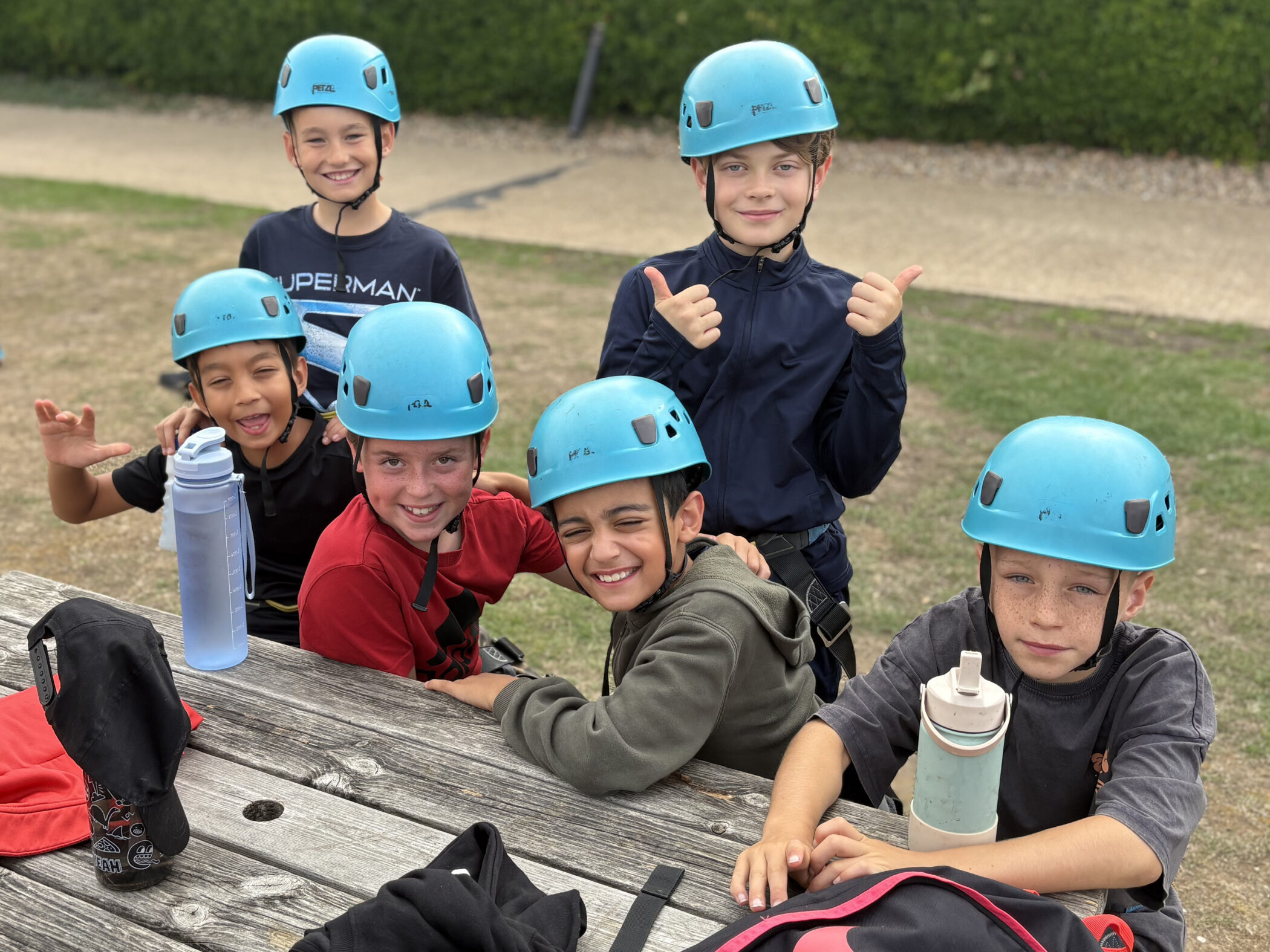 Six children wearing blue helmets sit at a picnic table outdoors, smiling and posing for the camera. Two water bottles and bags are on the table. The background includes grass and a dirt path.