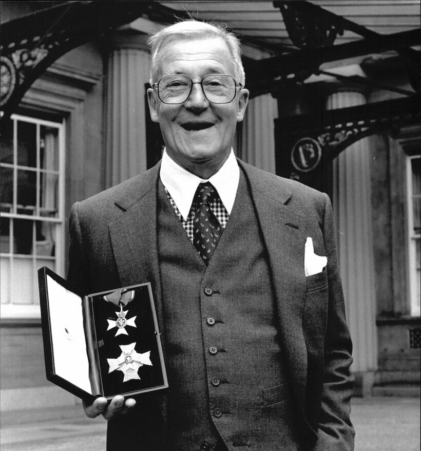 An older man in a suit smiles and holds an open box displaying medals or honours outside a formal building.