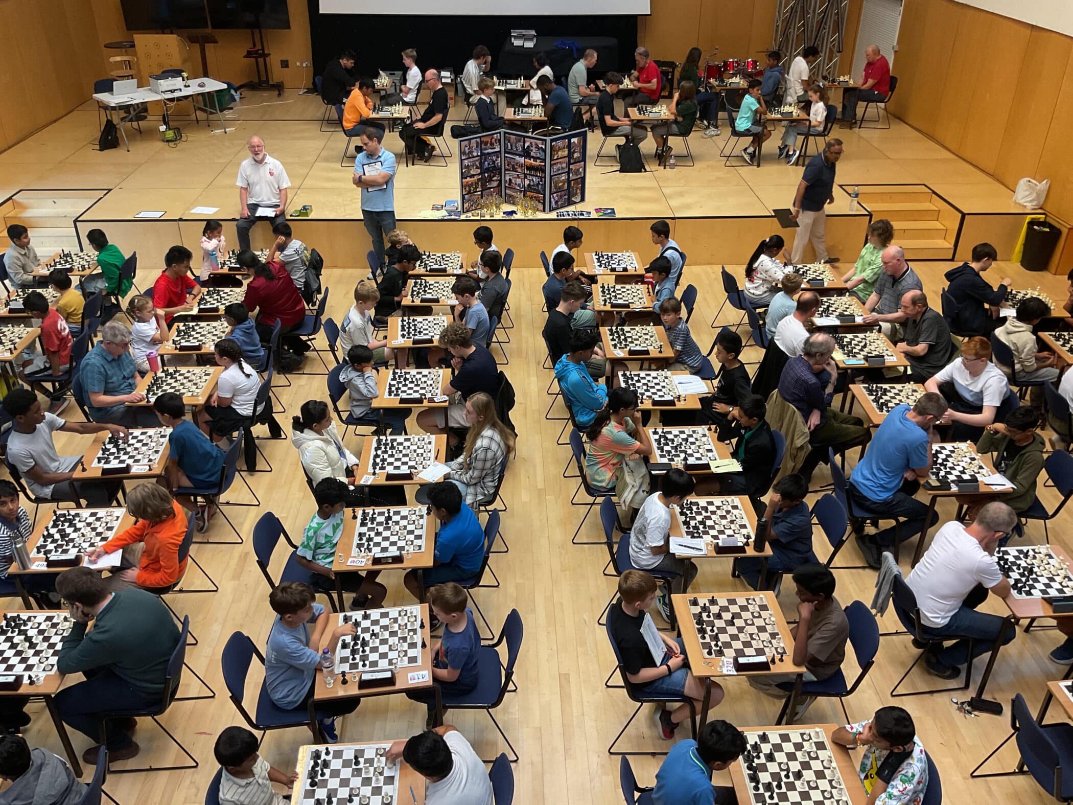 A large room filled with people of various ages playing chess at many tables, viewed from above. Spectators and organisers stand around, and a display board is set up in the background.