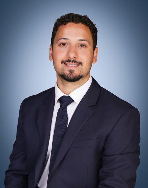 A man with short dark hair, a trimmed beard, and moustache is wearing a navy suit, white shirt, and dark tie, smiling slightly against a gradient blue background.