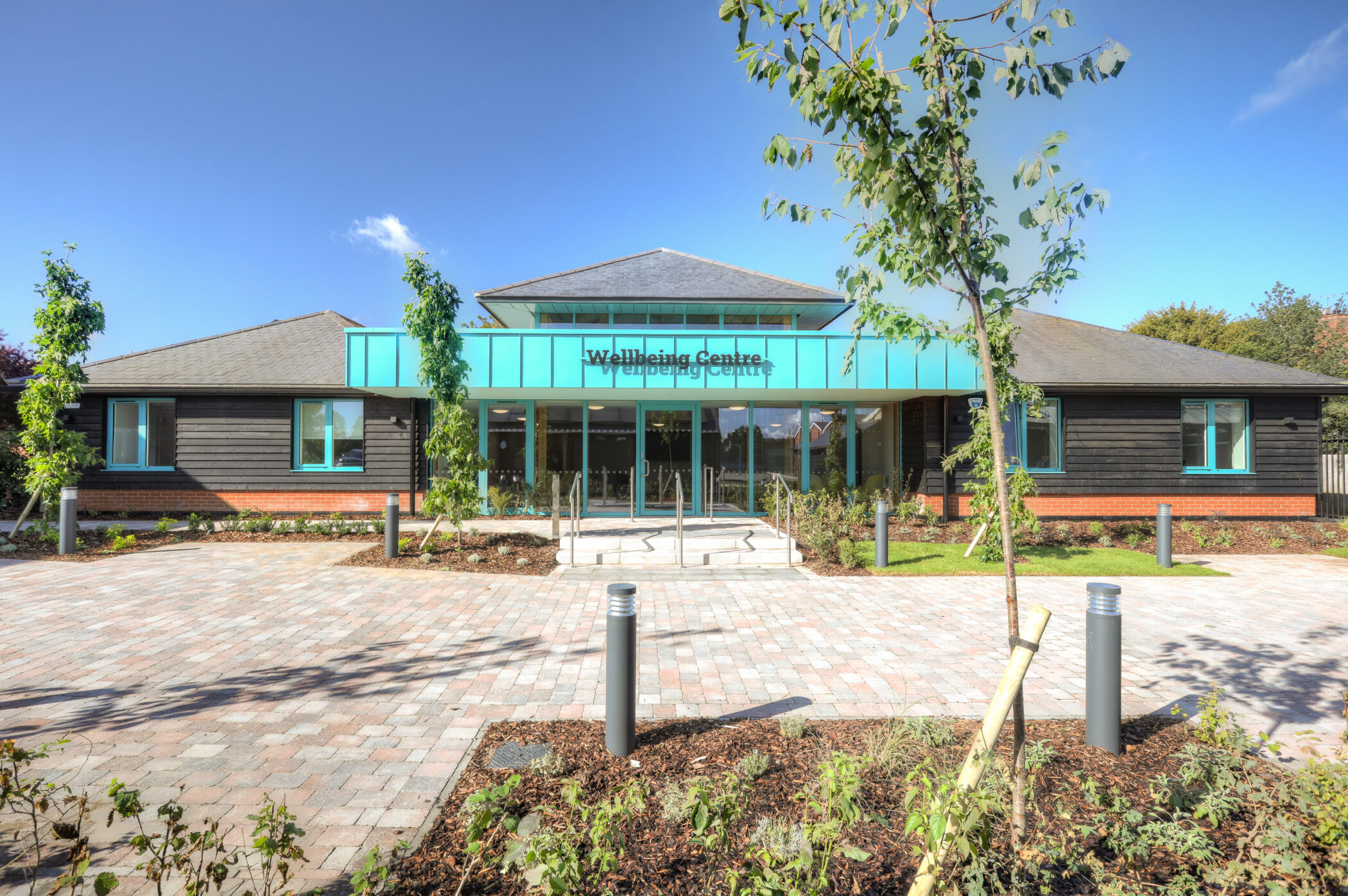 A modern, single-storey building labelled Wellbeing Centre stands as the focal point of this building, featuring a teal entrance and surrounded by young trees, landscaping, and a paved pathway under a clear blue sky.