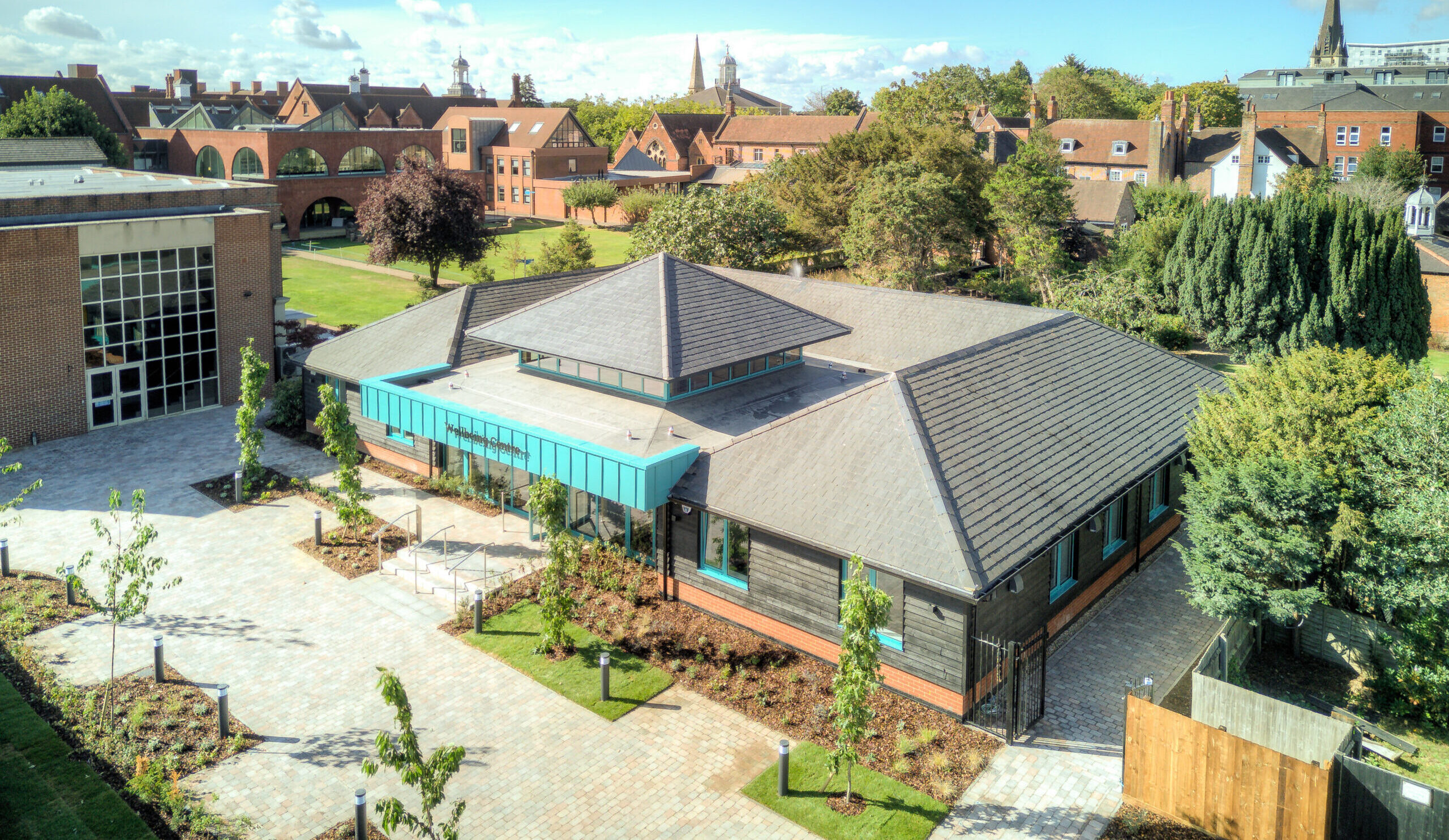 A modern, single-storey building with a grey roof is surrounded by trees and paved walkways. The Wellbeing Centre stands out against the backdrop of older brick buildings and church spires, all beneath a blue sky with scattered clouds.