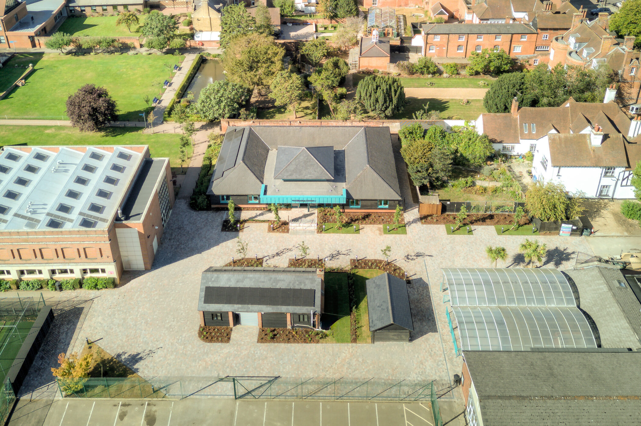 Aerial view of a school campus with several buildings, a greenhouse, tennis courts, trees and walkways on a sunny day, highlighting the new Wellbeing Centre at the heart of the grounds.