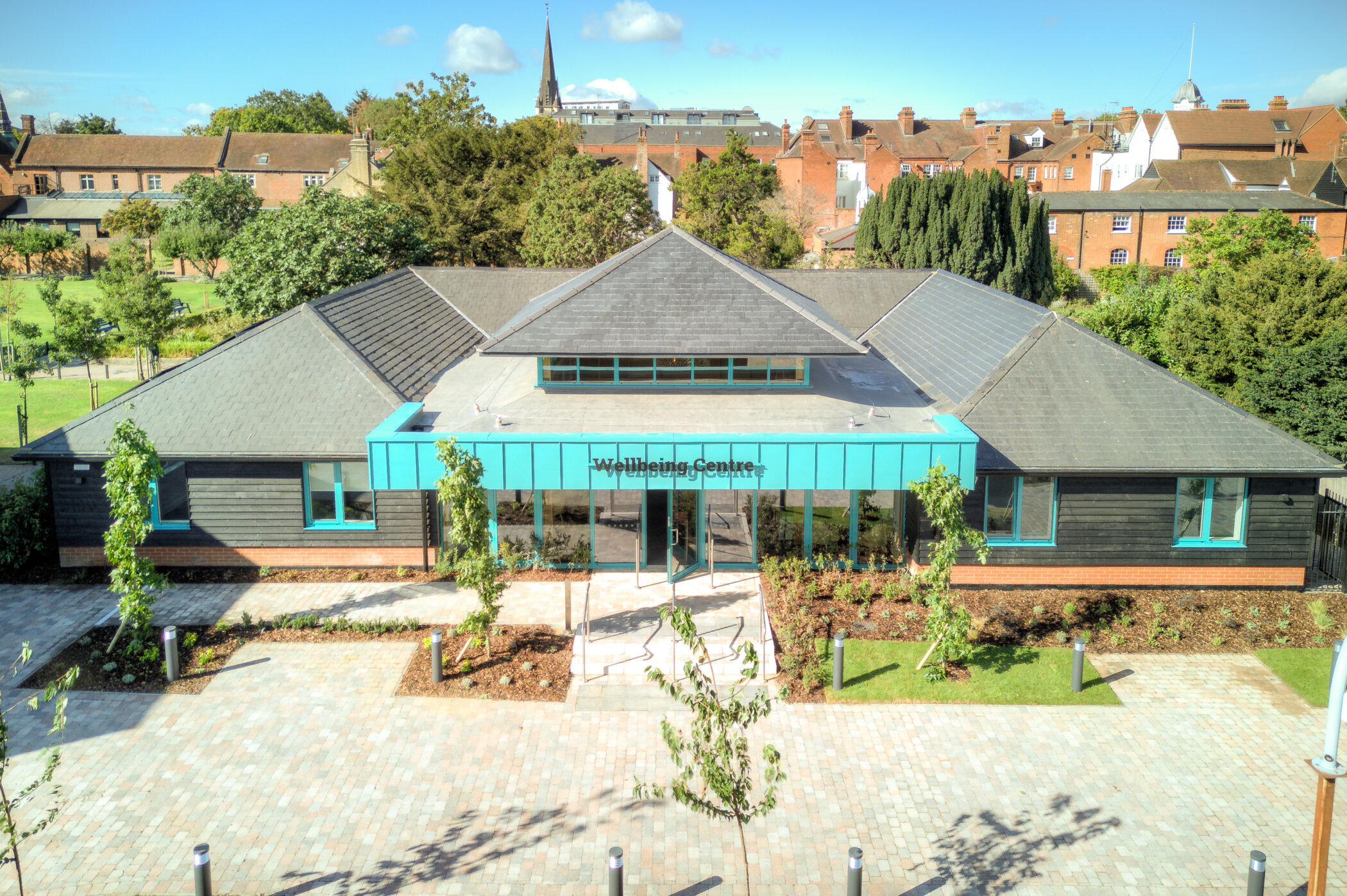 A modern building labelled Wellbeing Centre stands as a shining example of innovative Wellbeing at Brentwood School. The building features large windows and surrounded by trees, greenery, and a paved walkway under a blue sky with scattered clouds.