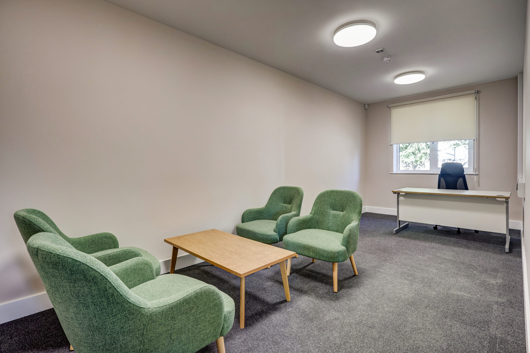 A modern office situated in the Wellbeing Centre features three green armchairs, a wooden coffee table, and a desk with a black chair by a window with a roller blind. The walls are plain and the room has grey carpet flooring.