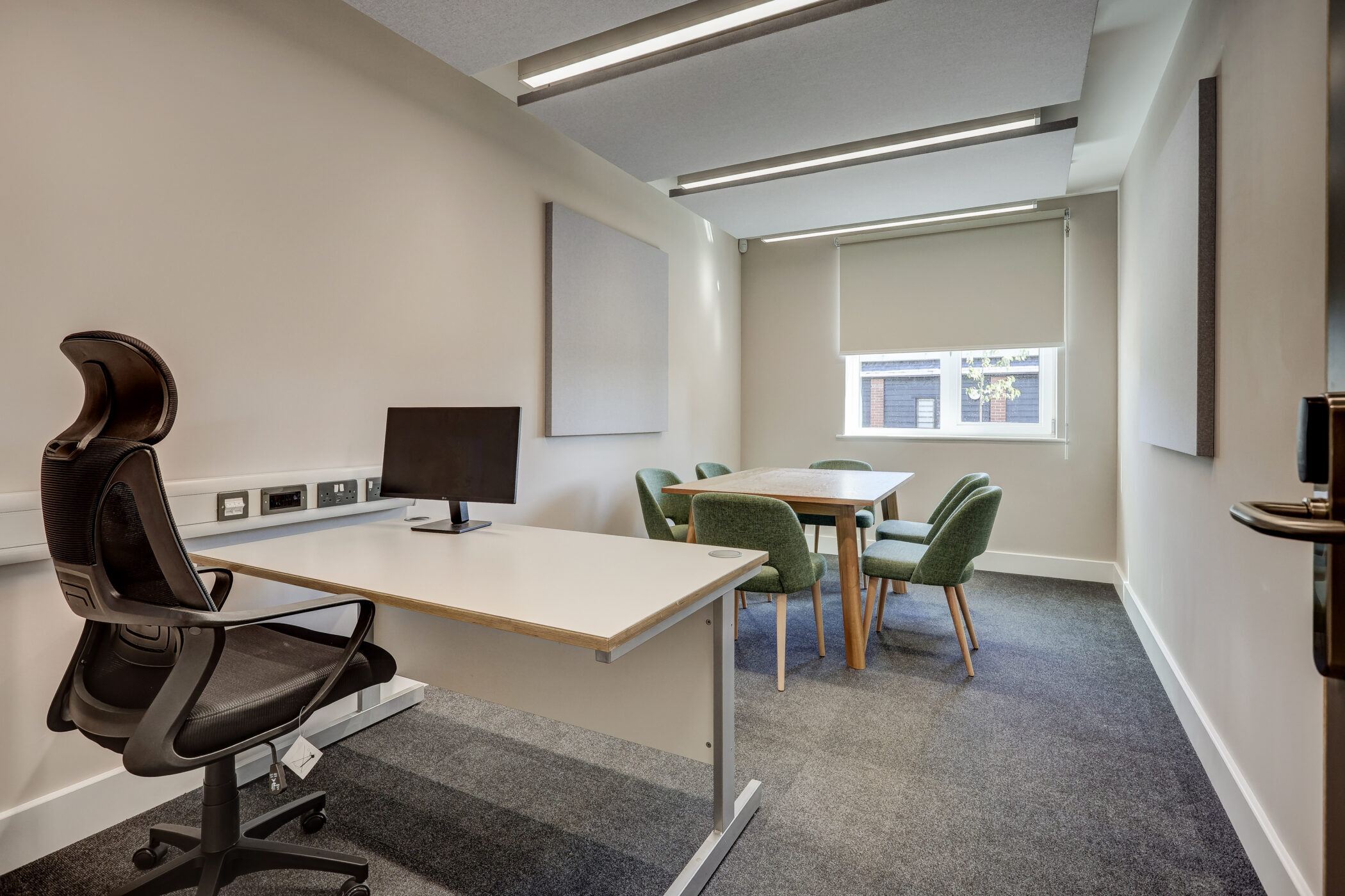 A modern office designed as part of the Wellbeing Centre, featuring a desk, computer, black chair, and a meeting table with four green chairs near a window with a roller blind. The room has light walls, carpeted floor, and ceiling lights.