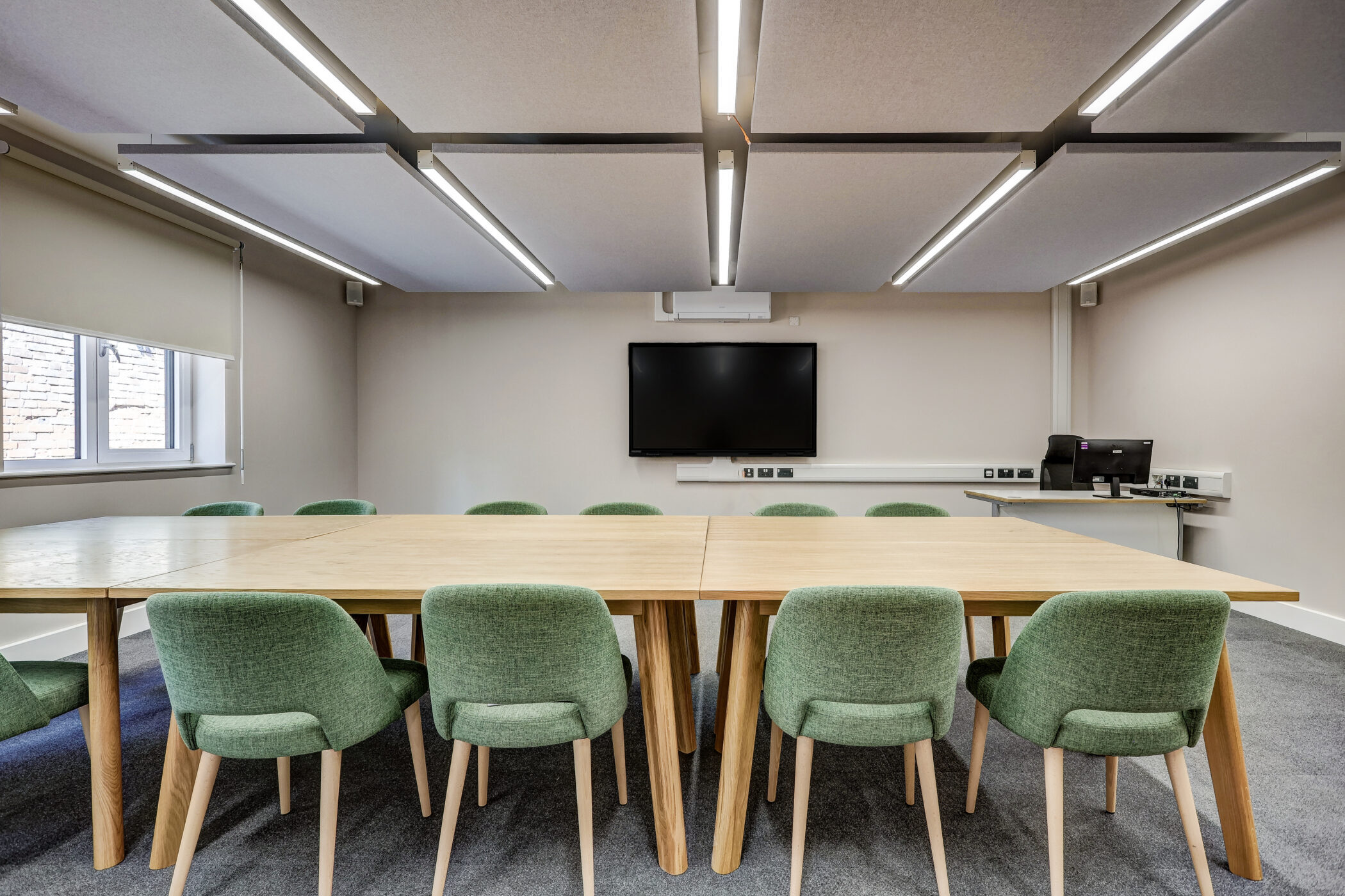 A modern studio room situated in the Wellbeing Centre, featuring a large wooden table, ten green upholstered chairs, a wall-mounted TV, and a computer on a side desk. The room has neutral walls, ceiling lights, and a window with blinds.