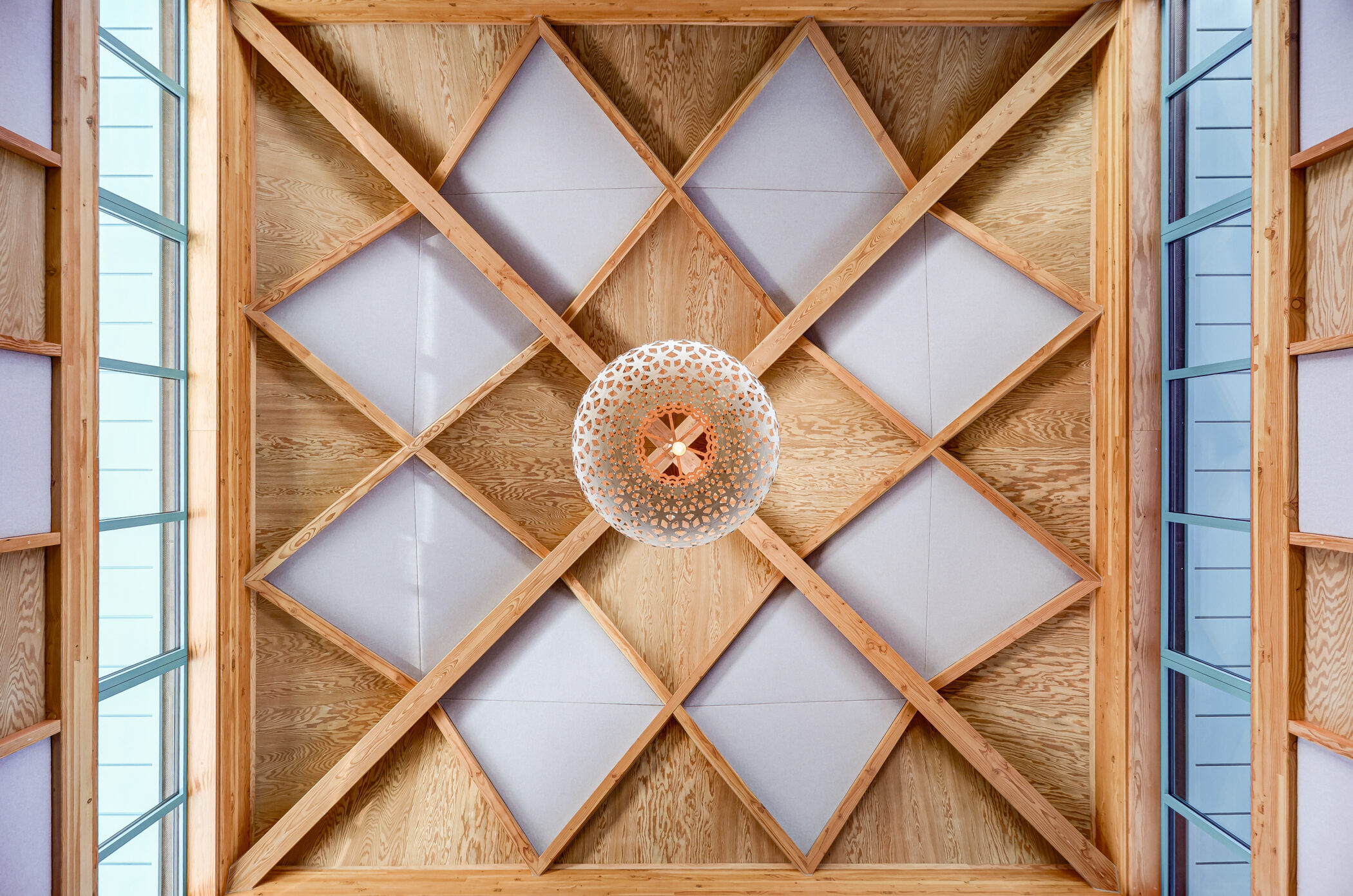 A geometric wooden ceiling with criss-crossed beams forming diamond shapes surrounds a round, decorative white light fitting in the centre, an architectural highlight perfect for the Wellbeing Centre. Tall windows frame the ceiling on both sides, inviting natural light and creating an atmosphere of tranquillity.