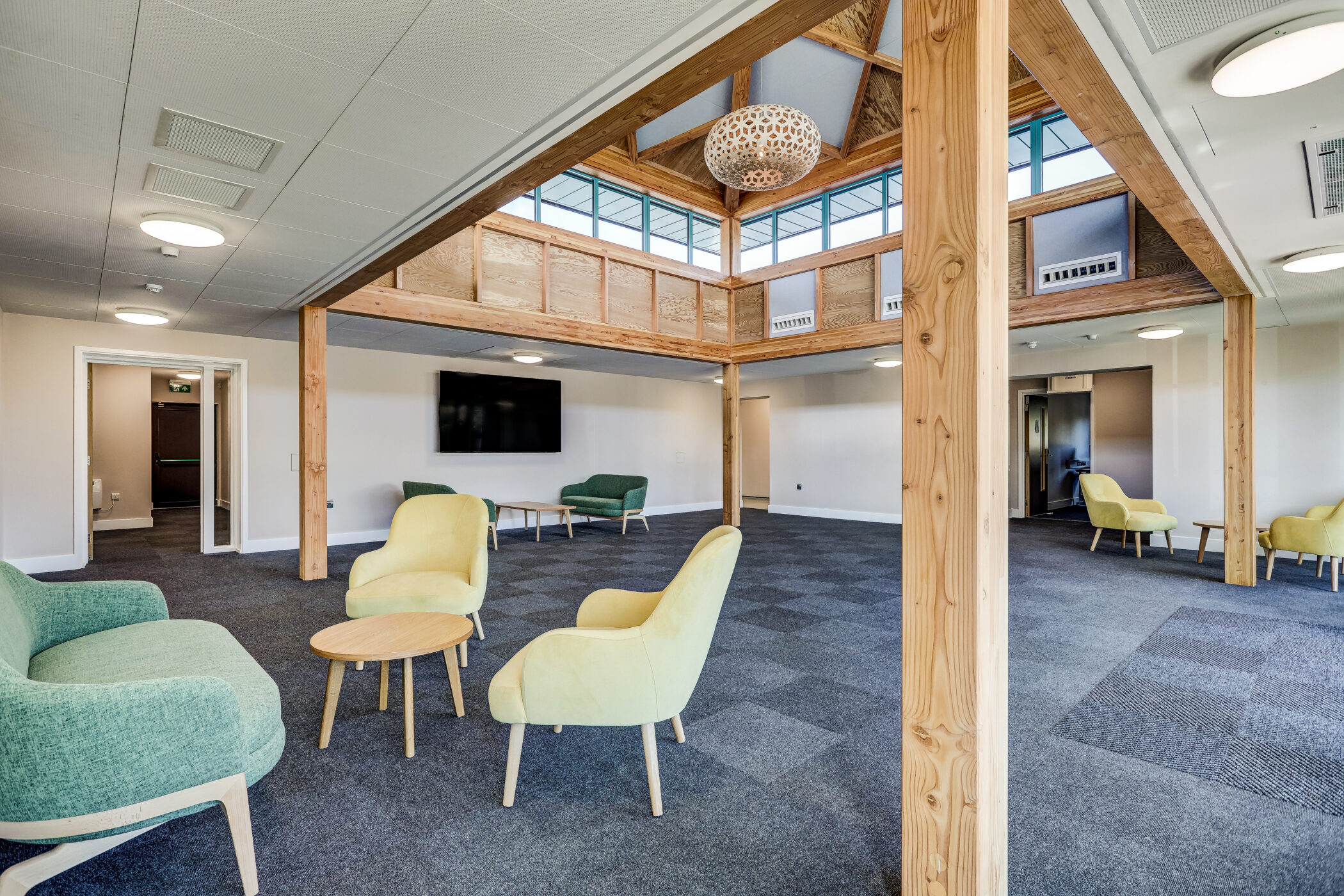 Spacious, modern lounge area with yellow and green chairs, small round tables, carpeted floor, wooden beams, large windows, and a central skylight, perfect for the Wellbeing Centre focused on relaxation and comfort.