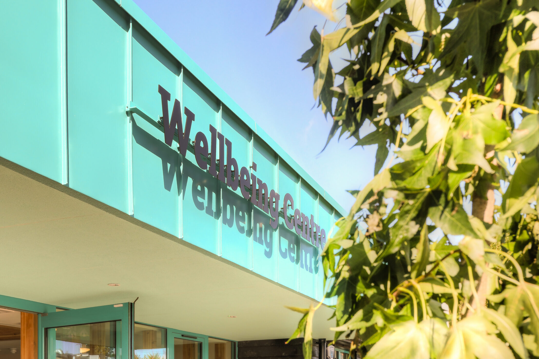 A teal building labelled Wellbeing Centre, is partially visible with green leaves in the foreground and a clear blue sky in the background.
