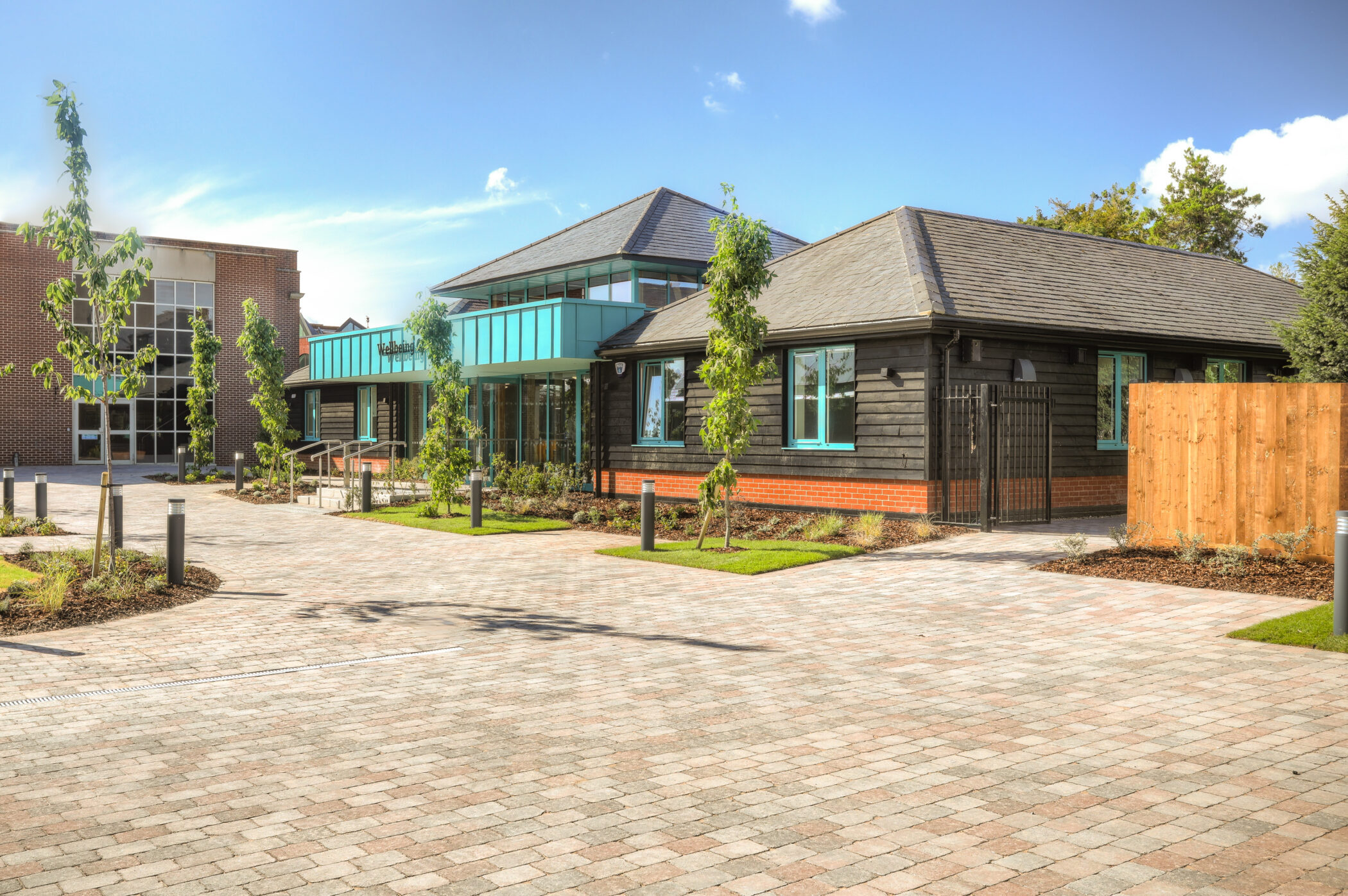 Two modern buildings with large windows and brick accents, part of the new Wellbeing Centre, are surrounded by fresh landscaping and young trees under a bright blue sky with scattered clouds. A paved walkway and driveway lie in the foreground.