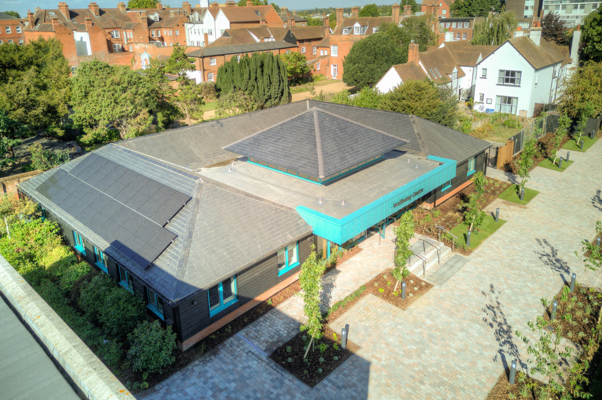 Aerial view of a modern, single-storey building with a grey roof and teal trim, surrounded by trees and brick pathways, situated in a residential neighbourhood, an ideal setting for a Wellbeing Centre.