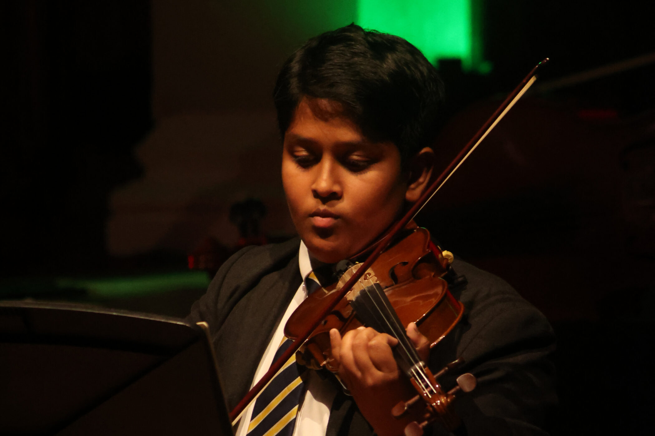 A person in a suit and striped tie is playing the violin on stage, reading sheet music. A green light glows in the background.