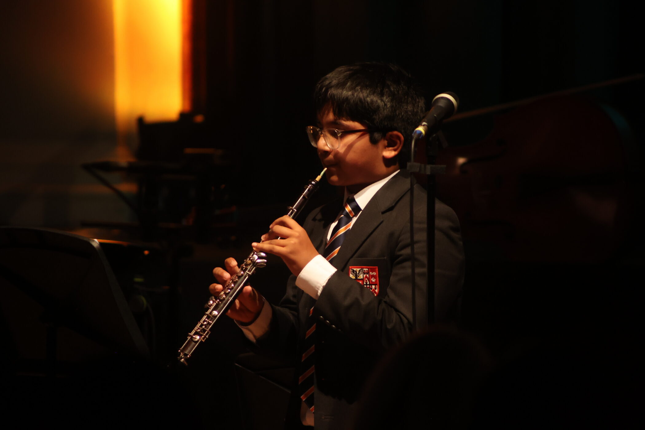 A young boy in a school uniform plays the clarinet on stage, standing in front of a microphone with warm lighting in the background.