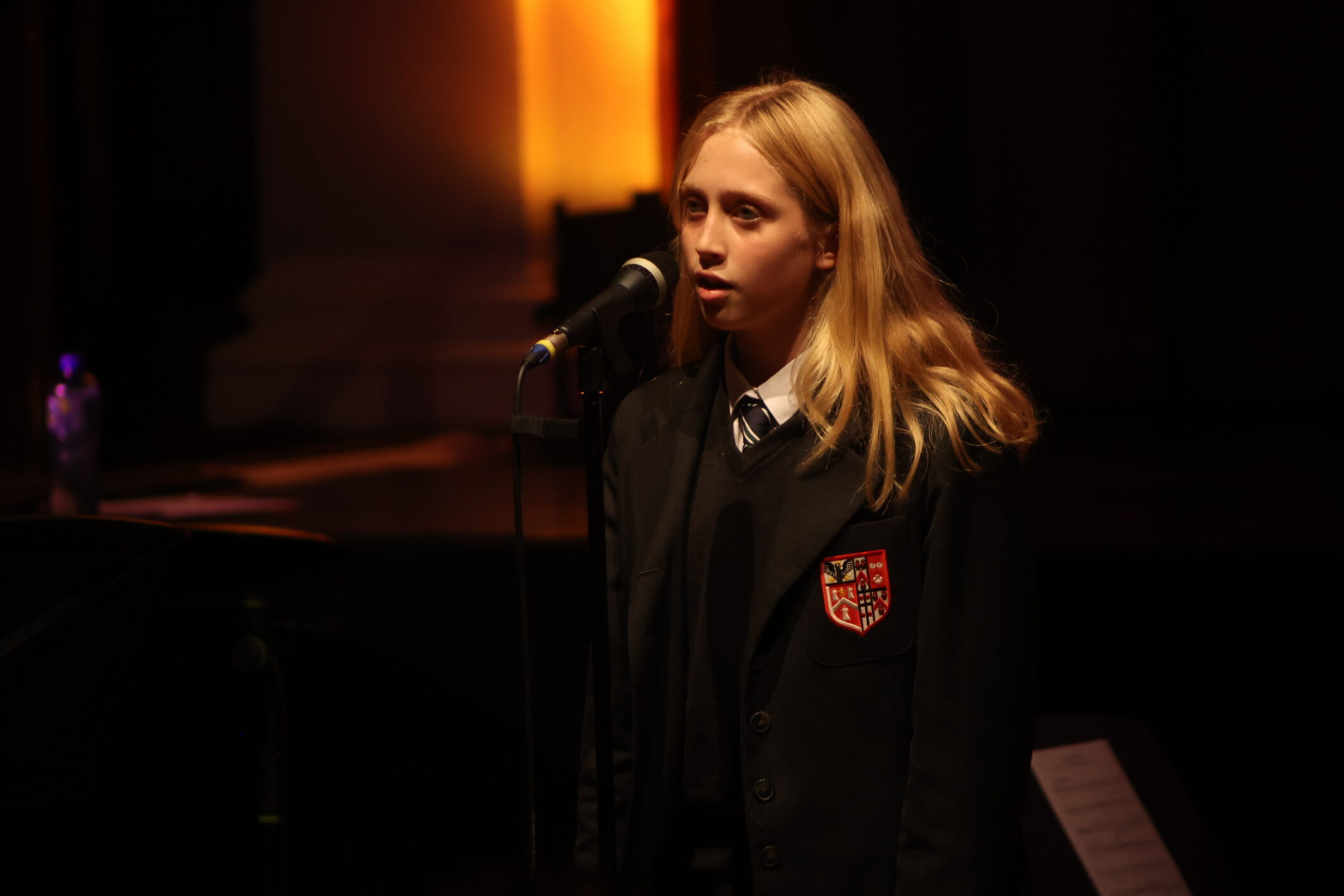 A blonde student in a dark blazer with a crest sings into a microphone on a dimly lit stage with warm lighting in the background.