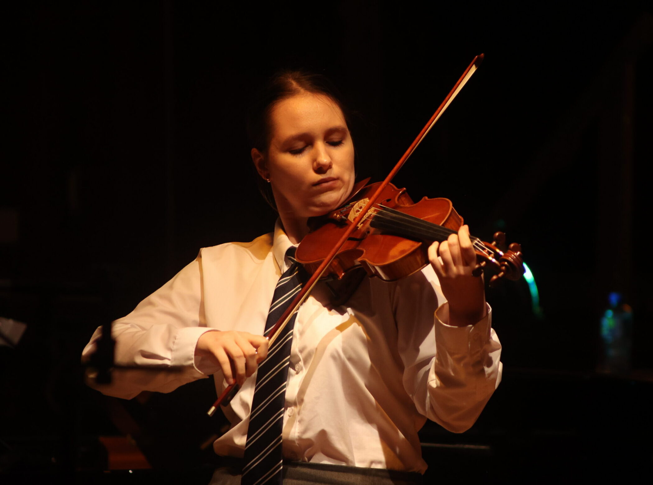A young person in a white shirt and tie plays the violin on stage at the Brentwood International Music Competition.