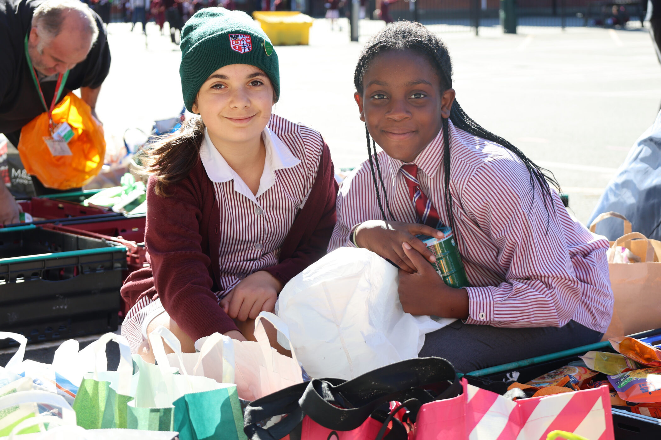 Two school-aged children in uniforms sit on the ground surrounded by bags of food donations.