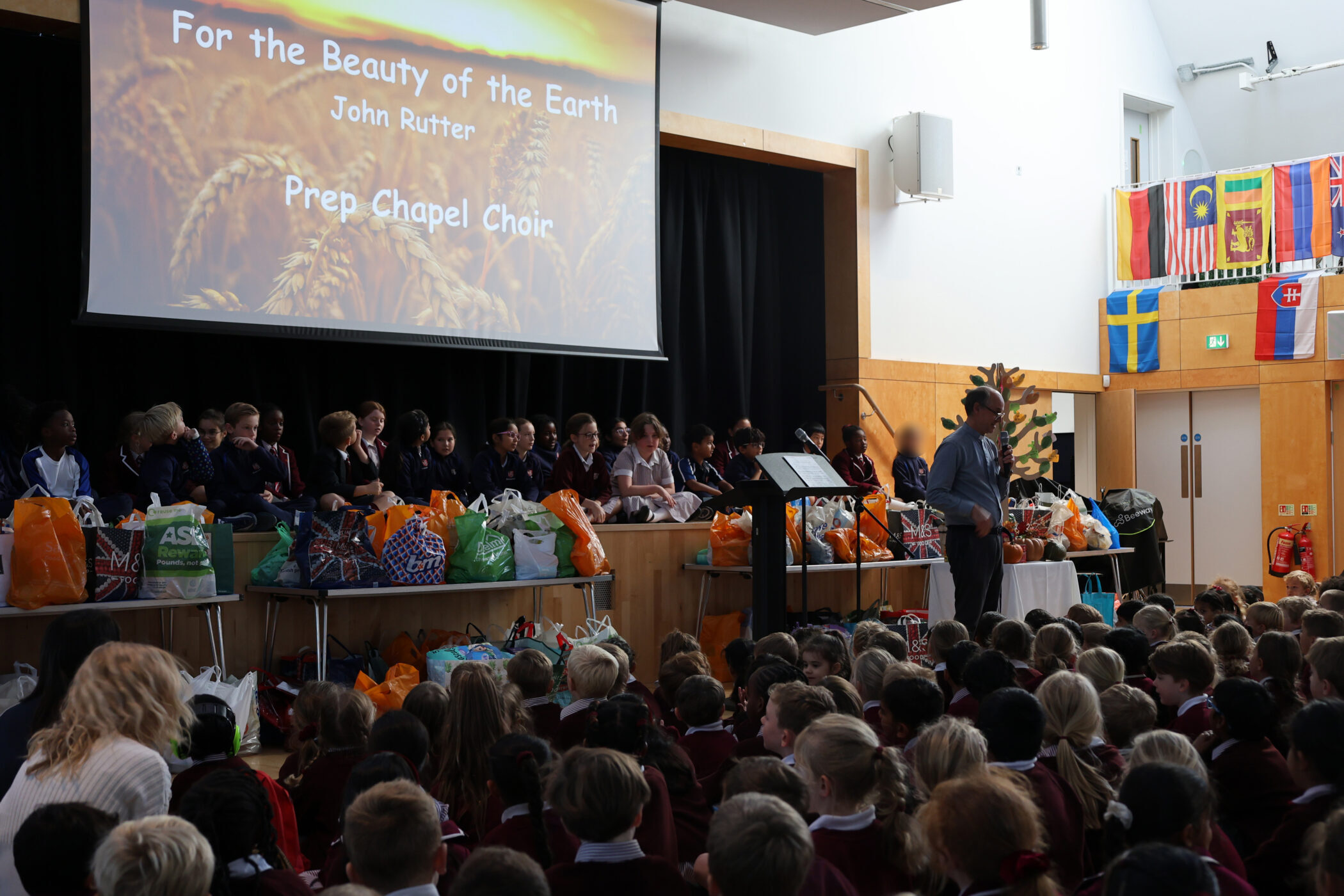 A school choir sits on a stage before a large screen displaying the title For the Beauty of the Earth whilst a man stands at a lectern addressing an audience of students.