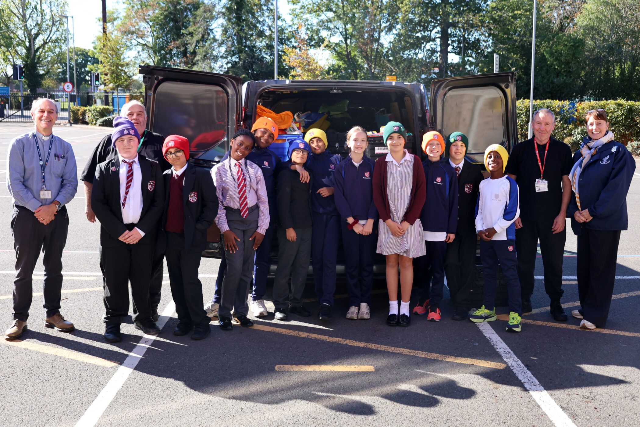 A group of students and adults stand in a car park in front of a van filled with supplies.