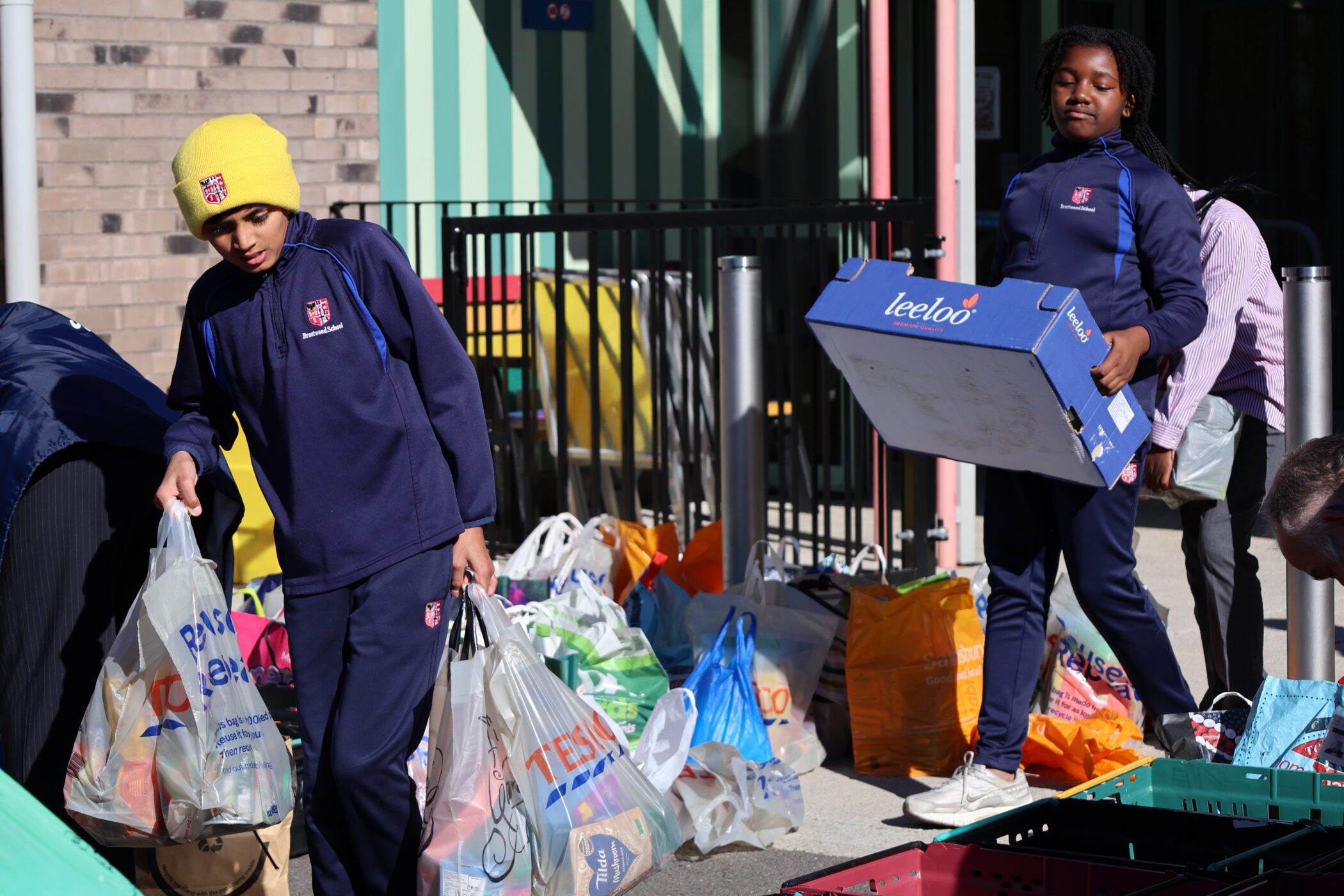 Two pupils in school uniforms carry bags and boxes of groceries outdoors.