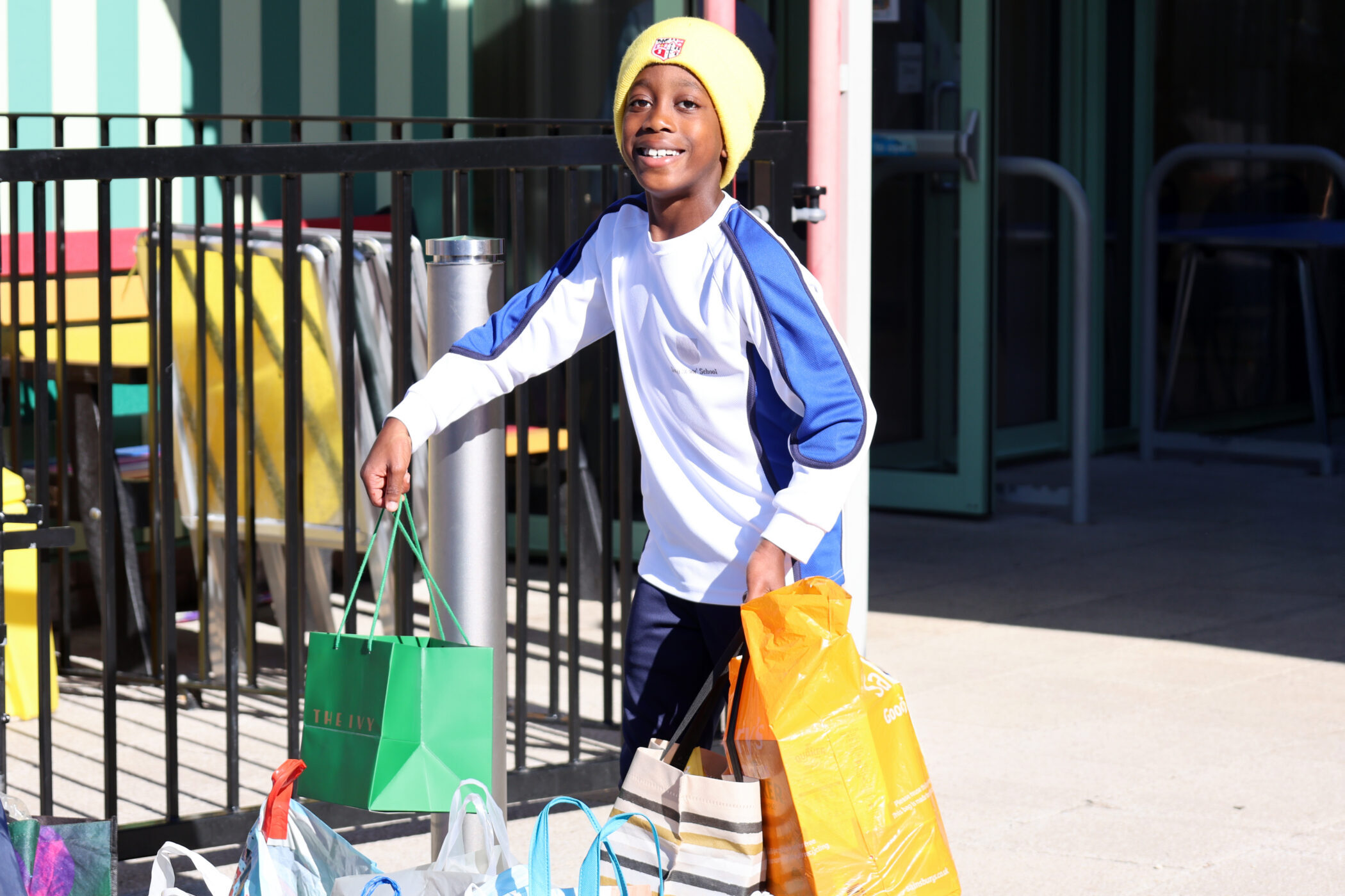 A smiling child wearing a yellow beanie and a white and blue long-sleeved shirt, carrying several shopping bags.