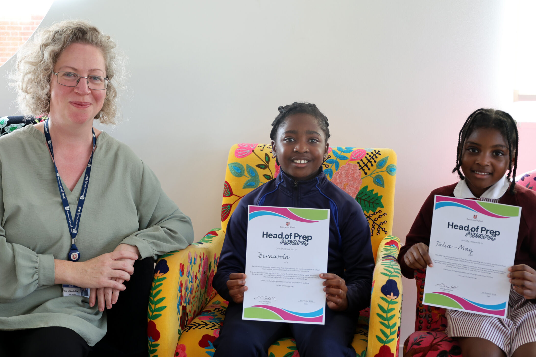A woman sits beside two smiling children in colourful chairs. The children hold Head of Prep Award certificates and look at the camera.