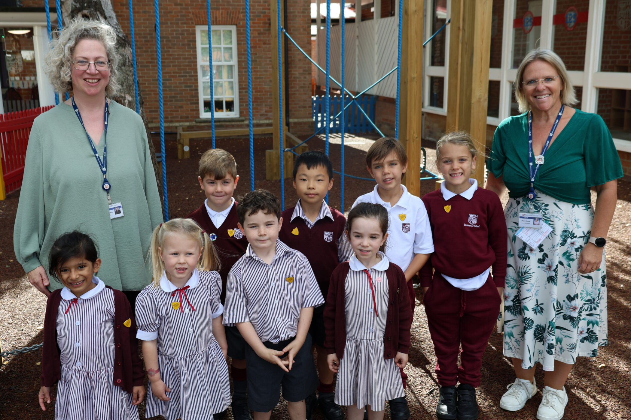 Two women and eight young children in school uniforms pose and smile outside on a playground with a brick building and a red fence in the background.
