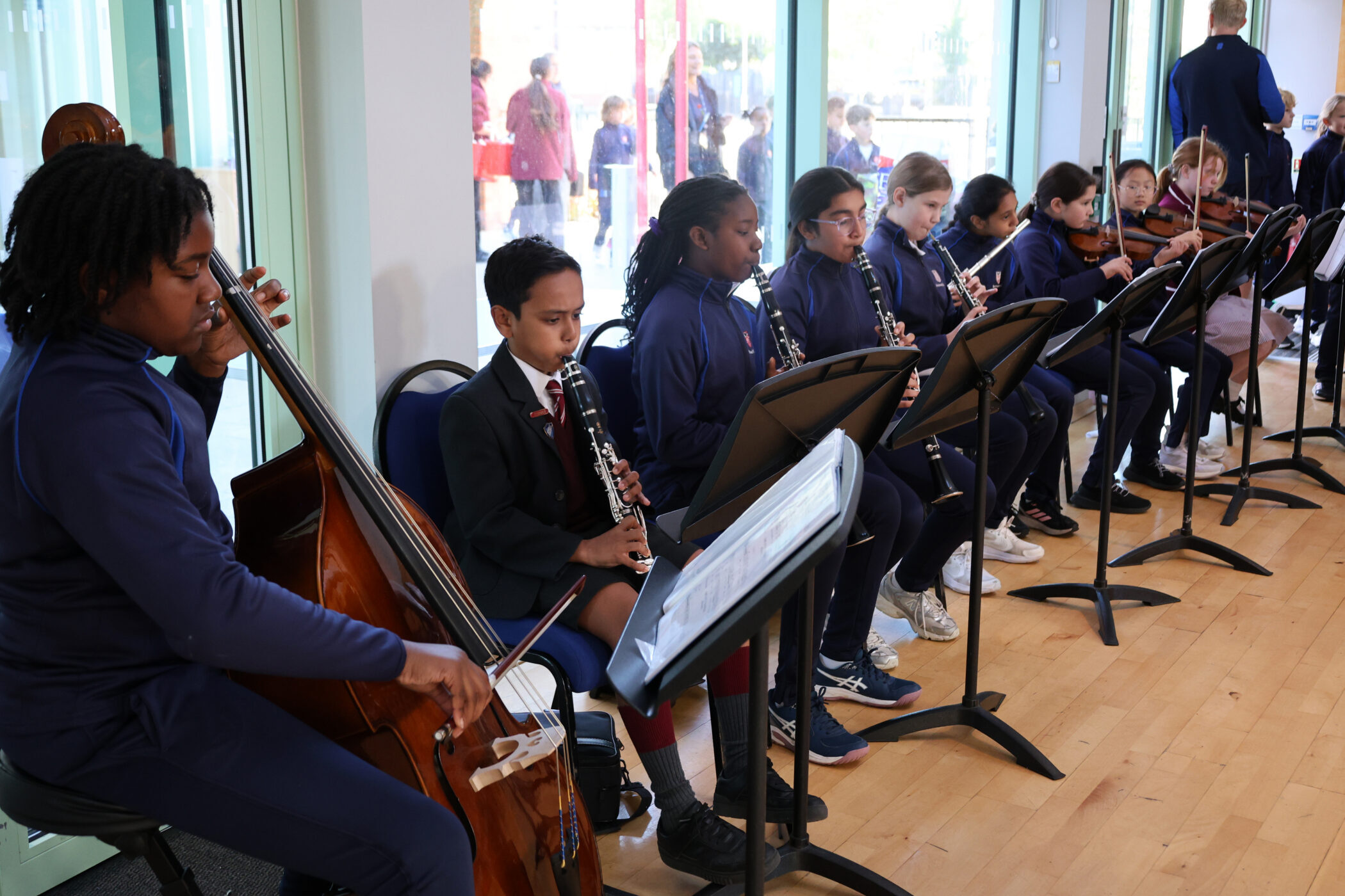 A group of students in matching navy uniforms playing musical instruments, including a double bass, clarinets, and violins, in a music practice room.