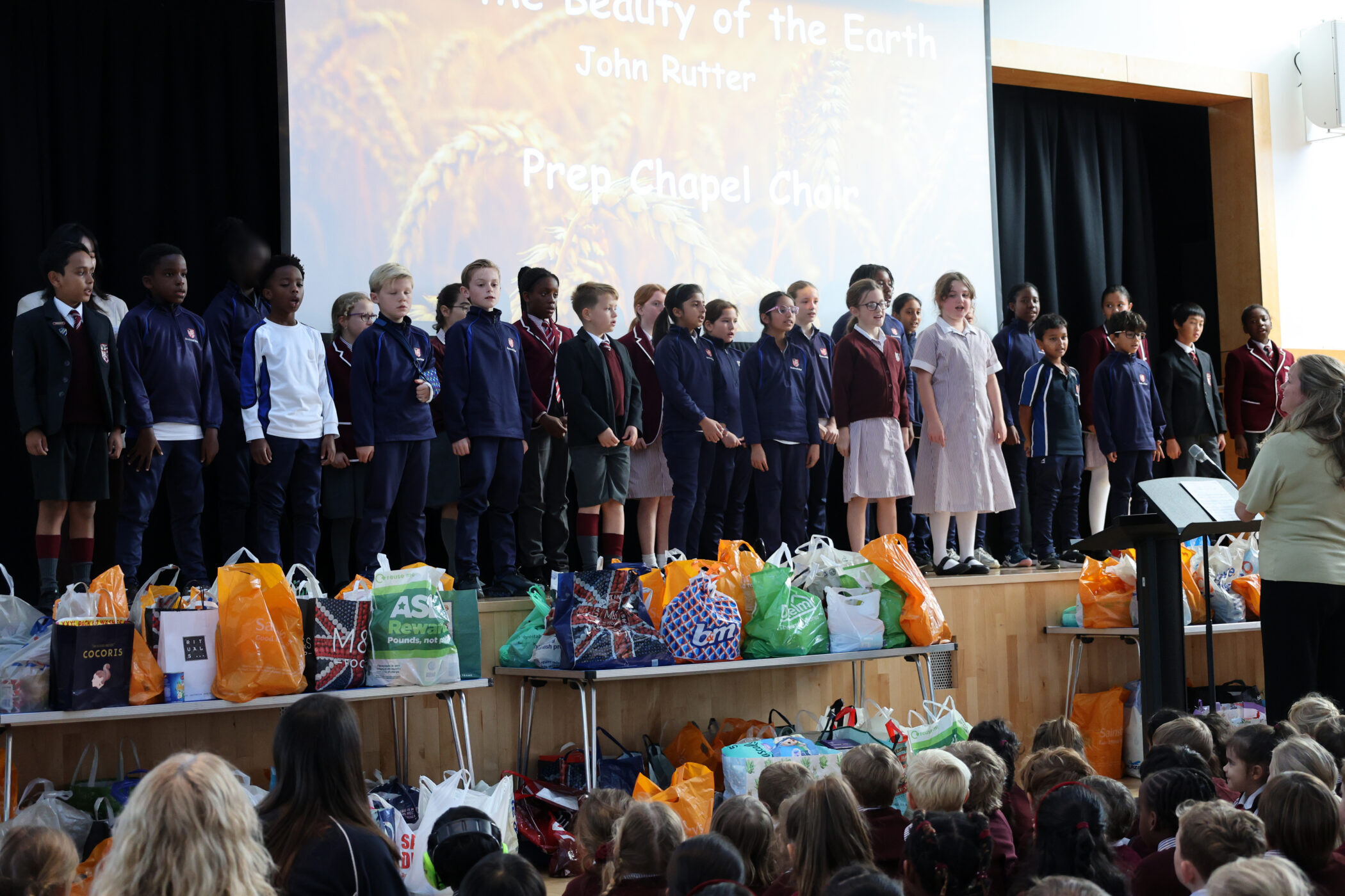 A choir of school children in uniforms stands on a stage in front of a projection screen, whilst a teacher at a music stand leads them before an audience of pupils.