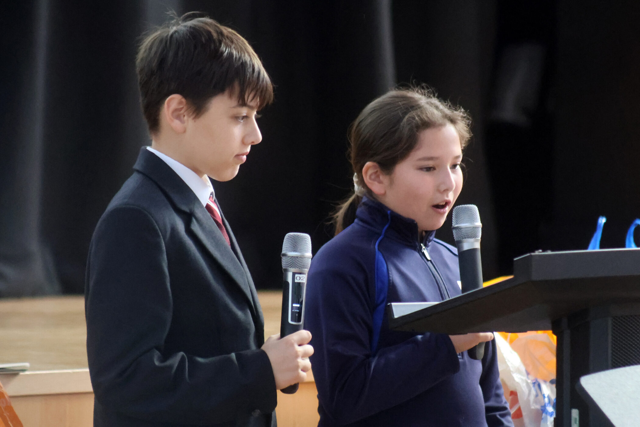 A boy in a suit and a girl in a tracksuit jacket stand at a lectern, each holding a microphone.