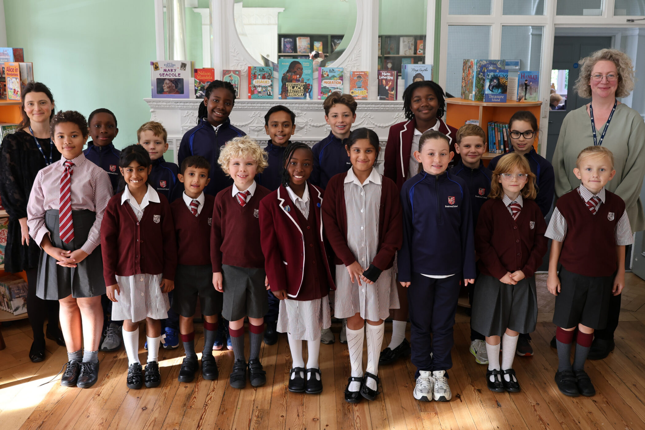 A large group of primary school children in uniform posing for a photo with their two teachers in a library.