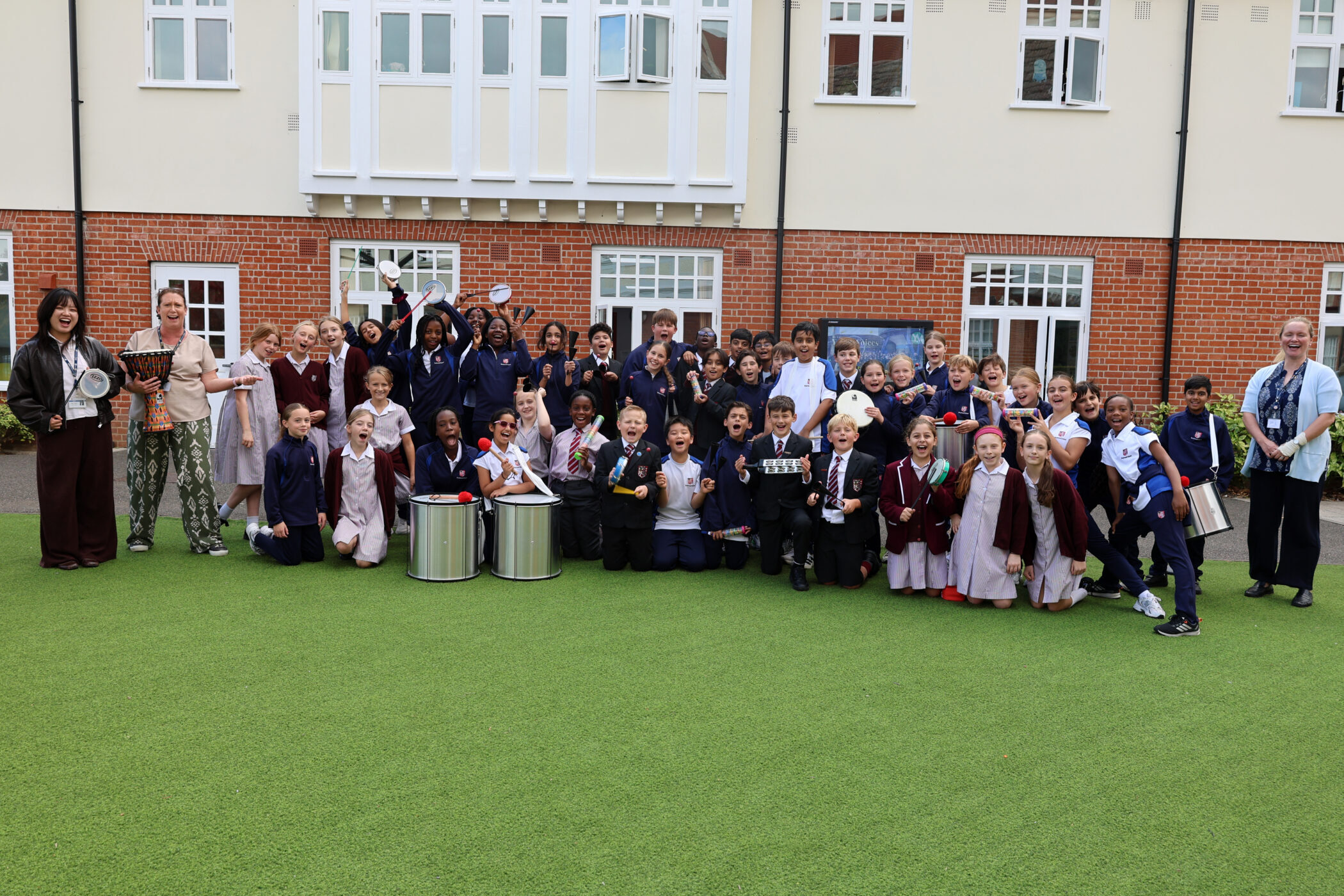 A large group of schoolchildren in uniforms, holding various percussion instruments, pose with two adult teachers on a grassy field in front of a brick building.