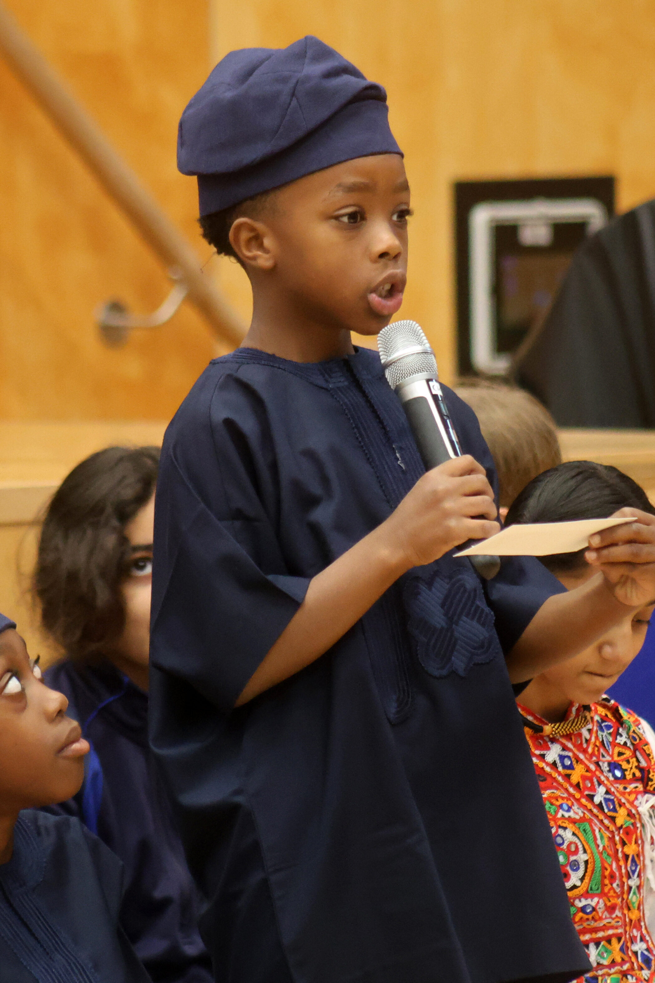 A young boy in a traditional dark blue outfit and cap speaks into a microphone whilst holding a small piece of paper.